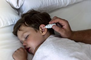 Father dribble medication into boy ear. Photo of a dad dripping ear drops to teenager son while sleeping in the comfortable bed at home. Ear drops. Close up man drips medicine into the ear of a sick boy.