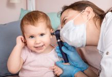 Pediatrician examines ear of baby girl at home during coronavirus COVID-19 pandemic quarantine. Doctor using otoscope (auriscope) to check ear canal and eardrum membrane of a child