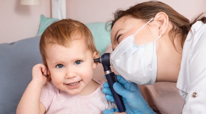 Pediatrician examines ear of baby girl at home during coronavirus COVID-19 pandemic quarantine. Doctor using otoscope (auriscope) to check ear canal and eardrum membrane of a child
