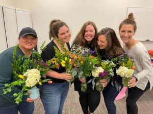 A group of women, joined together at a mom's group, is standing together and smiling with their flowers