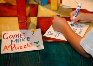 A child lettering a sign with a blue marker next to a sign that says "Come make a puzzle!: