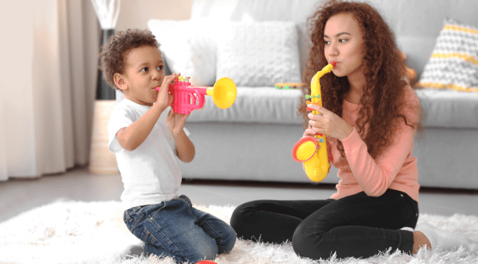 Female babysitter with long brown curly hair playing a yellow toy saxophone on the floor of a home next to a biracial male child playing a toy horn.