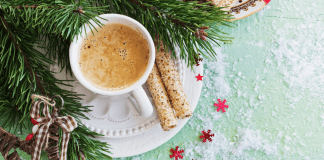 Cup of coffee with cream in a white mug on a fancy saucer sitting on a table with a mint green tablecloth dusted in sugar and the plate has evergreen leaves on it and cinnamon sticks for story on holiday coffee drinks